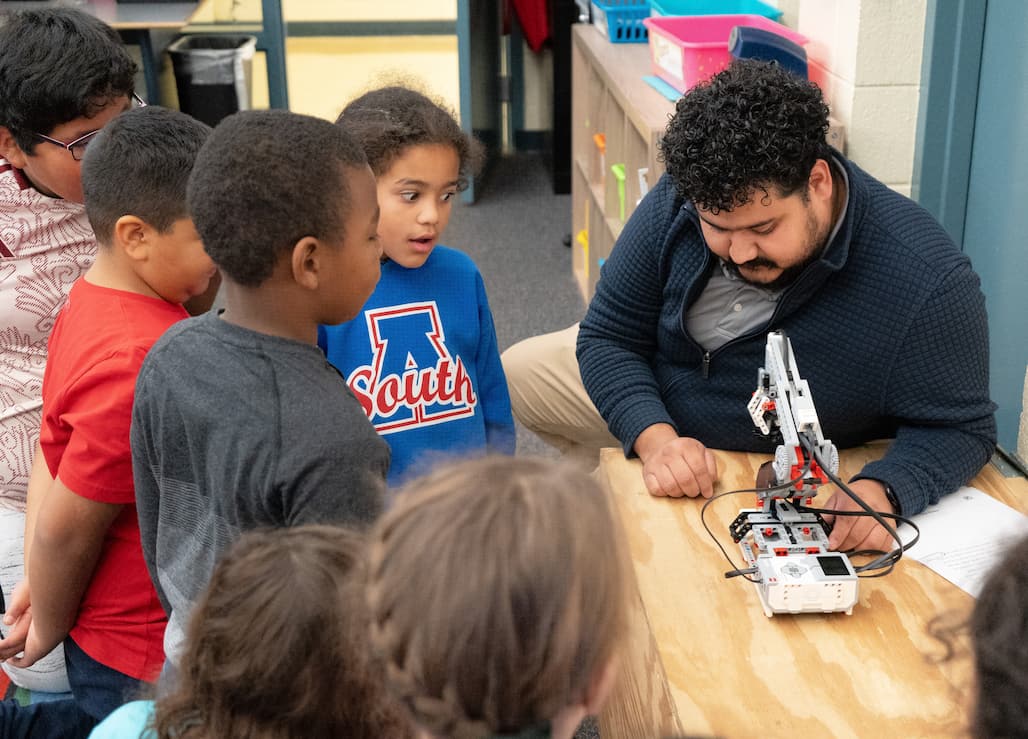 Photo of a person demonstrating a robotic arm made of Legos while a group of students watches.