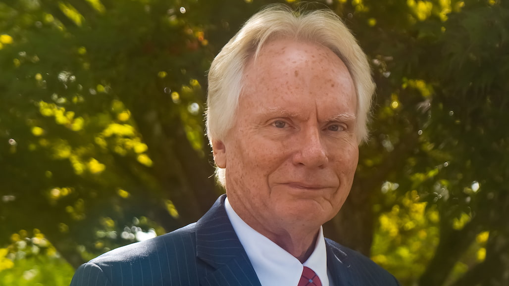 Photo of a man wearing a suit and standing in front of a maple tree.