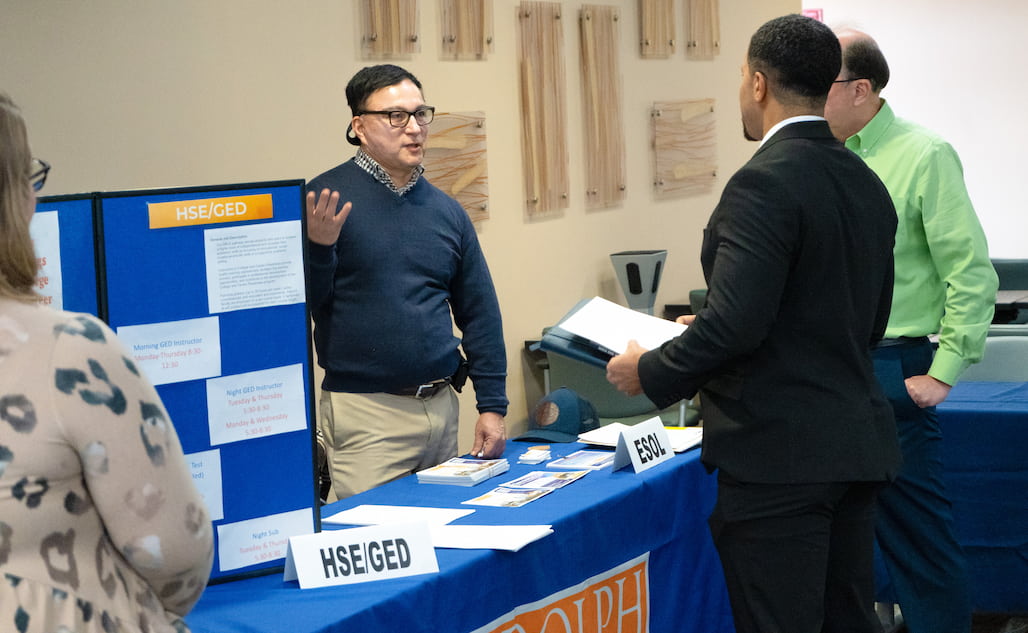 Photo of a person standing at a table speaking to another person.