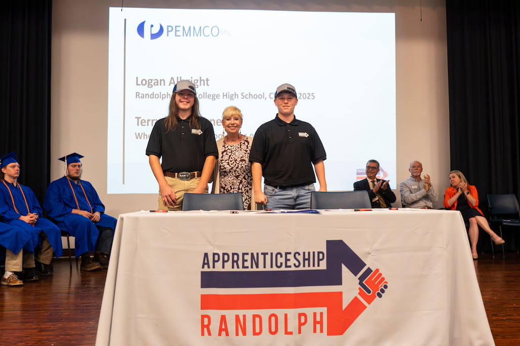 Two Apprenticeship Randolph signees stand in front of a table with the program’s logo, joined by a woman at center stage. A presentation slide behind them shows their names and school affiliations. Seated graduates in blue caps and gowns and community leaders in business attire applaud in the background.