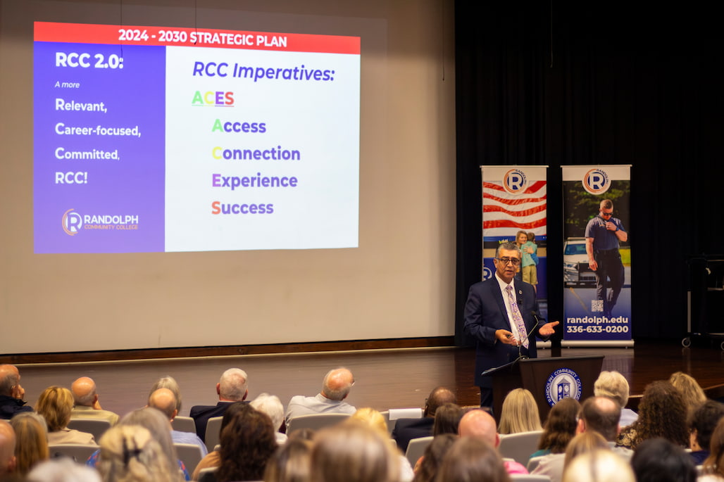 Dr. Shah Ardalan, President/CEO of Randolph Community College, speaks at a podium in front of a large audience. A presentation slide on the screen shows RCC’s 2024–2030 Strategic Plan, highlighting "RCC 2.0: A more Relevant, Career-focused, Committed RCC!" and the RCC Imperatives: ACES — Access, Connection, Experience, Success. Two RCC banners stand on the stage behind him.